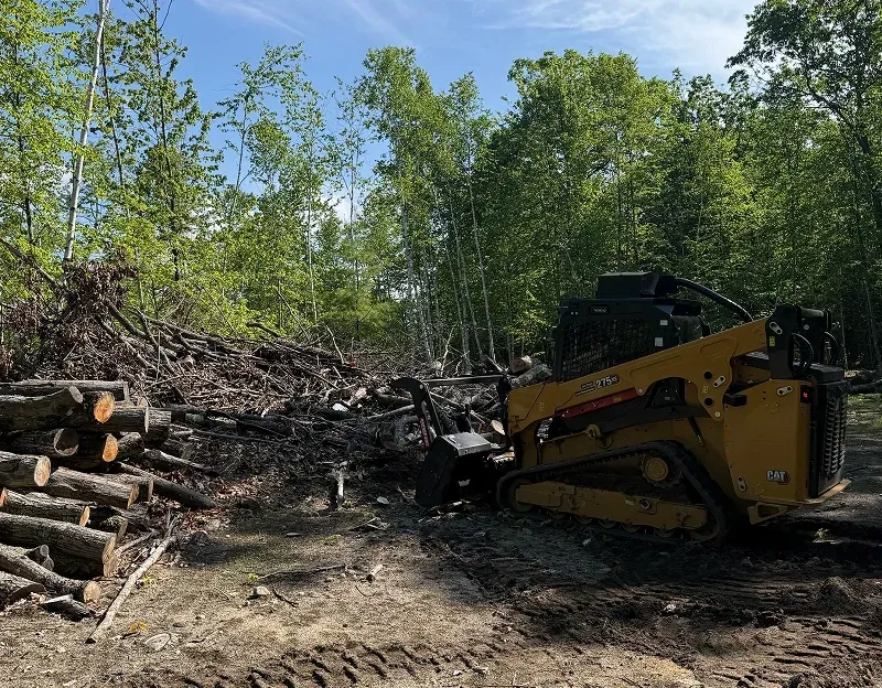Track loader with a forestry mulcher beside cut logs and a brush pile